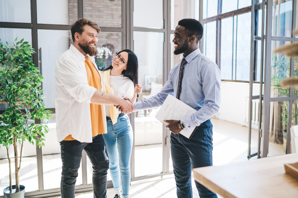 Real estate broker with contract and smiling couple sealing deal of apartment purchase by shaking hands